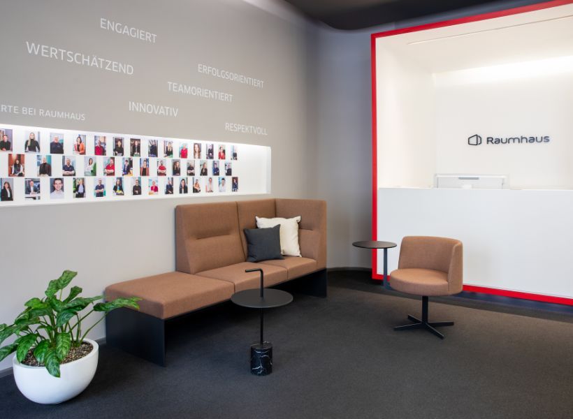 Wartebereich mit Sofa, Sessel und Beistelltischen vor einer Empfangstheke; an der Wand befinden sich Mitarbeiterfotos und Unternehmenswerte.