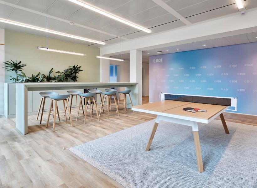 Workcafé mit hoher Theke, Barhockern, Pflanzen und Tischtennisplatte als kombinierter Bereich für Austausch, Pausen und informelle Zusammenarbeit.