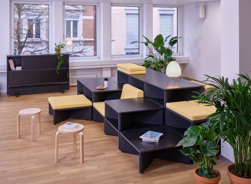 Modulare Sitzlandschaft mit gepolsterten Elementen, kleinen Tischen und Pflanzen in einer ruhigen Bürozone für informellen Austausch und konzentriertes Arbeiten.