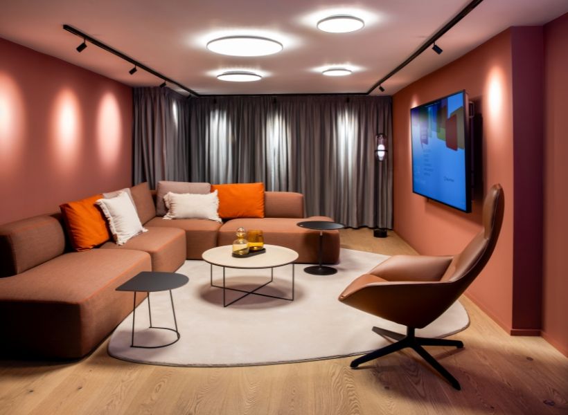 Loungeartiger Büroraum mit modularer Sofalandschaft in warmen Rosé‑ und Orangetönen, runden Beistelltischen, Teppich und Wanddisplay, stimmungsvoll beleuchtet durch Deckenleuchten.