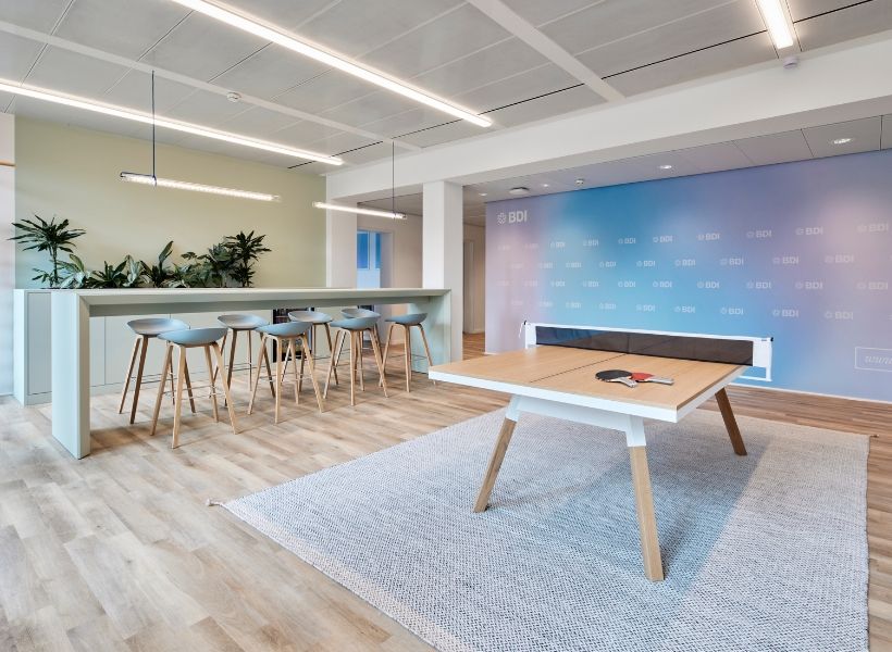 Heller Marktplatz mit hohem Stehtisch, Barhockern, Tischtennisplatte und Pflanzenakzenten für Teams zur Regeneration.