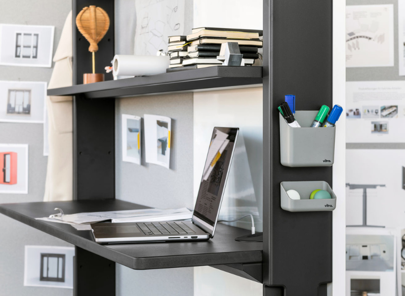 Detailaufnahme eines Arbeitsplatzes mit Laptop, schwarzer Ablagefläche, offenem Regalsystem, Notizmaterial und kleinen Organisationsbehältern.