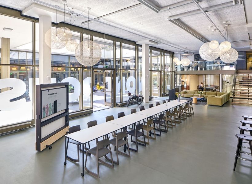 Großer offener Büroraum mit langen Arbeitstischen, mobiler Präsentations‑Stellwand und Lounge‑Bereich für Zusammenarbeit.