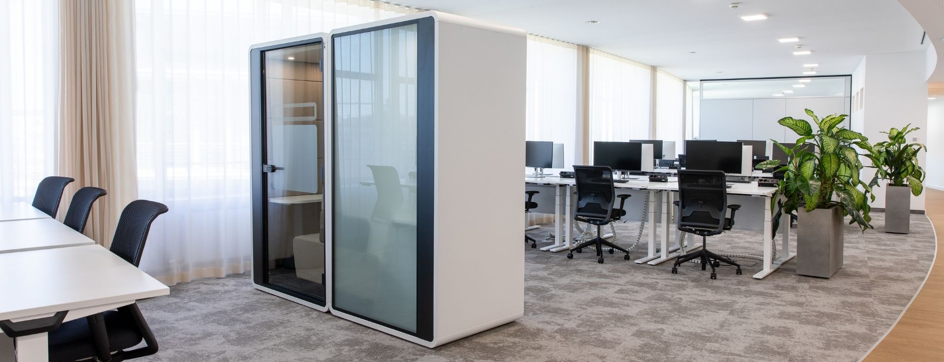 Telefonbox im offenen Bürobereich, positioniert neben ergonomischen Arbeitsplätzen und großen Fenstern mit heller Vorhanggestaltung.