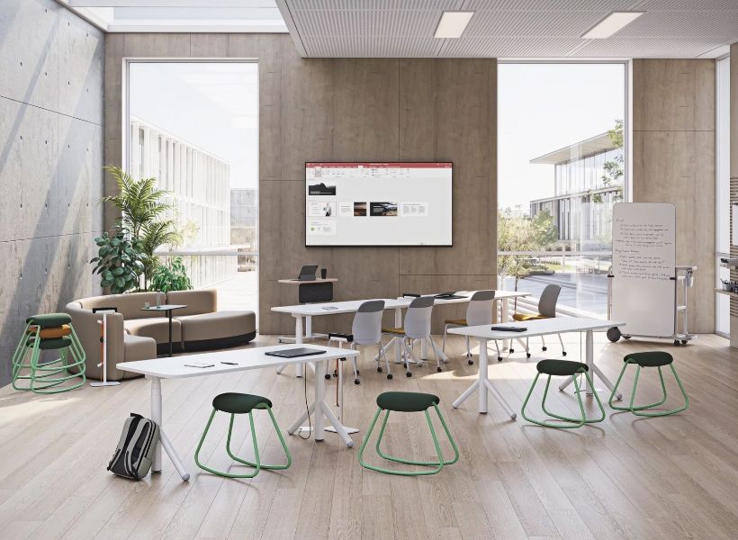 Flexibler Teamarbeitsbereich mit mobilen Tischen, Hockern, Whiteboard und Bildschirm für Workshops oder Besprechungen vor großen Fensterflächen.