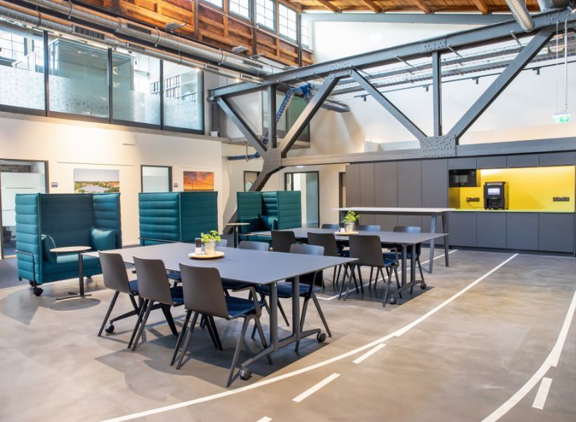 Großzügiges Workcafé in einer offenen Bürohalle mit Gemeinschaftstischen, geschützten Sitznischen und integrierter Kaffeeküche für Austausch und informelles Arbeiten.