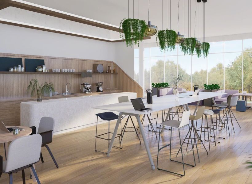Workcafé mit Küchenzeile, großem Hochtisch, Sitzgelegenheiten und begrünten Pendelleuchten vor bodentiefen Fenstern für Austausch und mobiles Arbeiten.