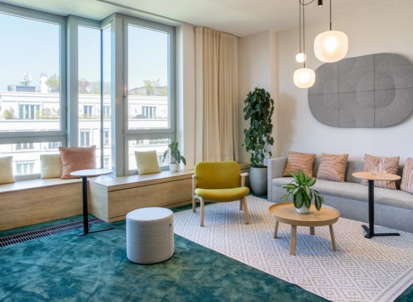 Heller Aufenthaltsbereich im Büro mit Sofa, Sesseln und Beistelltischen in Beige‑, Grün‑ und Gelbtönen, großem Fensterfront, Teppich, Pflanzen und Pendelleuchten in ruhiger Atmosphäre.