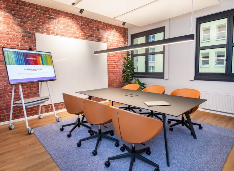 Moderner Besprechungsraum mit Backsteinwand, schwarzem Tisch, orangen Stühlen, großem Display und akzentuierter Deckenbeleuchtung.