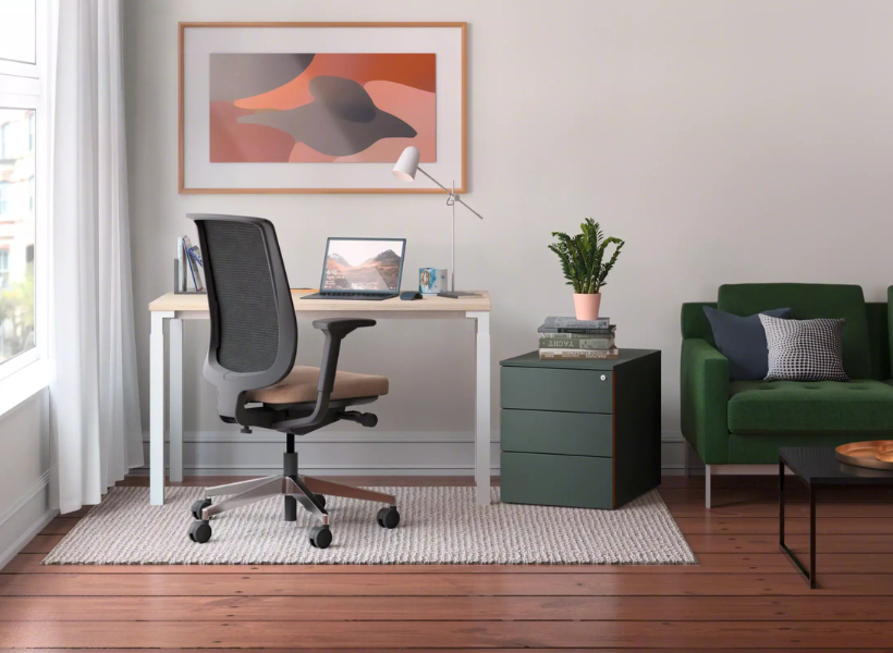 Helles Homeoffice mit schlichtem Schreibtisch, ergonomischem Drehstuhl, Tischleuchte und Laptop, ergänzt durch ein grünes Sofa und eine kleine Kommode mit Pflanze.