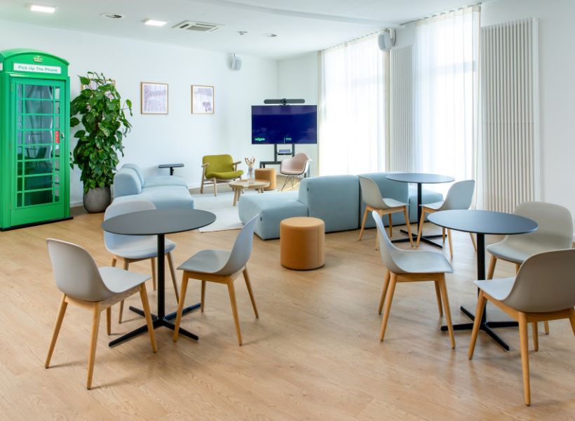 Offen gestaltetes Workcafé mit Café-Tischen, Lounge-Möbeln, Videokonferenzbildschirm und Besprechungskabine für informelle Treffen und Zusammenarbeit.