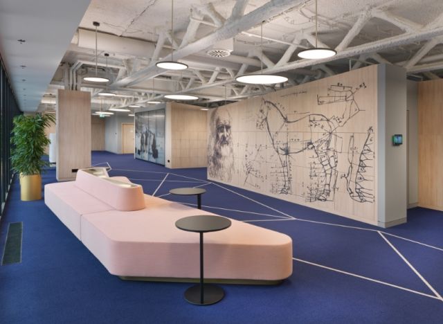 Offener Kommunikationsbereich mit blauer Teppichfläche, rosa Sitzinsel und großformatiger Wandgrafik, ergänzt durch runde Beistelltische und moderne Deckenbeleuchtung.