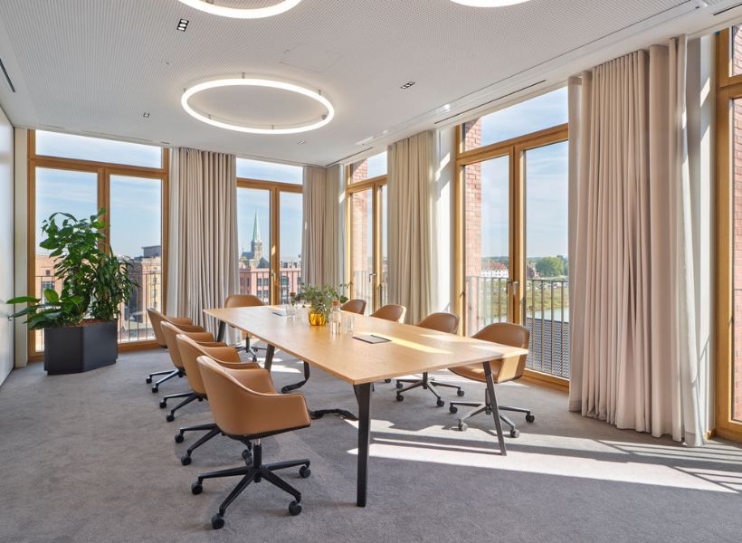 Besprechungsraum mit großem Tisch, großzügigem Tageslicht, ringförmigen Deckenleuchten und warmen Materialien für eine ausgewogene Arbeitsplatzbeleuchtung.