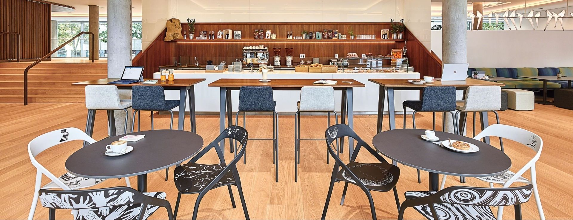 Großzügiges Workcafé mit Kaffeebar, Stehtischen, Sitzgruppen und Arbeitsplätzen mit Laptops als Kombination aus Begegnungs- und Arbeitsbereich im Büro.