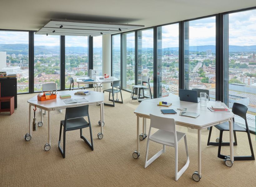 Arbeitsbereich mit mobilen Schreibtischen, Stühlen und großzügigen Fensterfronten in offener Raumgestaltung.
