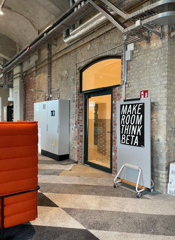Schild mit dem Schriftzug „MAKE ROOM THINK BETA“ vor einer Tür im Vitra BETA Event Space; sichtbare Industriearchitektur mit freiliegenden Leitungen und Backsteinwänden.