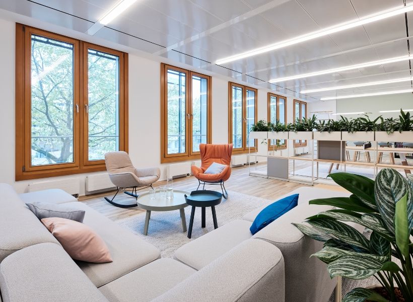 Offene Loungezone mit Sofas, Sesseln und Beistelltischen vor bodentiefen Fenstern, ergänzt durch Pflanzen und helle Holzdetails.