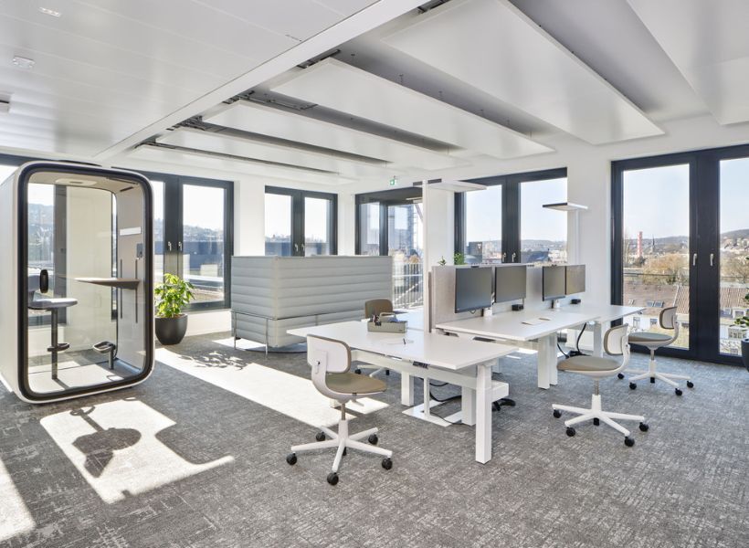 Offene Bürofläche mit höhenverstellbaren Schreibtischen, Bildschirmarbeitsplätzen, Besprechungskabine und gepolstertem Soft-Seating.