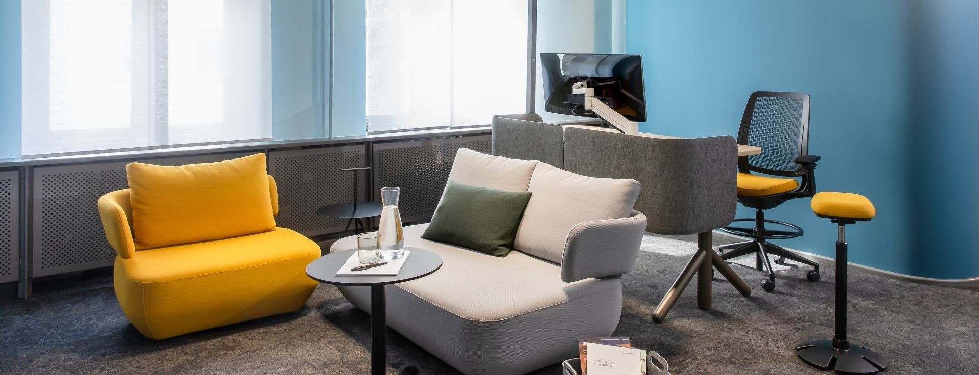 Moderner Büroraum mit Lounge‑Sesseln und Sofa in Grau‑ und Gelbtönen, kleinem Rundtisch, Teppichboden und Arbeitsplatz im Hintergrund vor blauer Akzentwand.