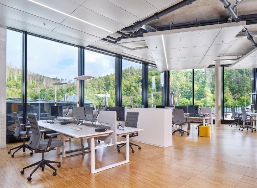Open-Space-Büro mit großzügigem Tageslicht, höhenverstellbaren Arbeitsplätzen und zonierter Struktur für konzentriertes Arbeiten und Austausch.
