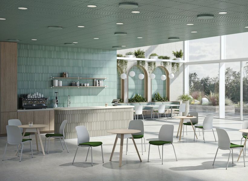 Modernes Workcafé mit Kaffeetheke, kleinen Sitzgruppen und begrünten Nischen vor großen Fensterflächen für Pausen und informellen Austausch im Büro.