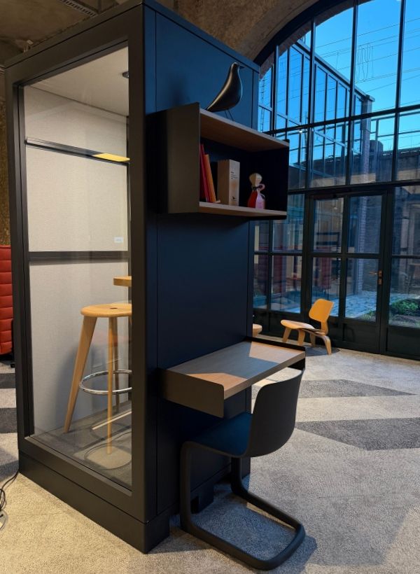 Akustisch abgeschirmte Arbeitskabine mit integriertem Schreibtisch und Regal vor großflächigen Industrie-Fenstern.