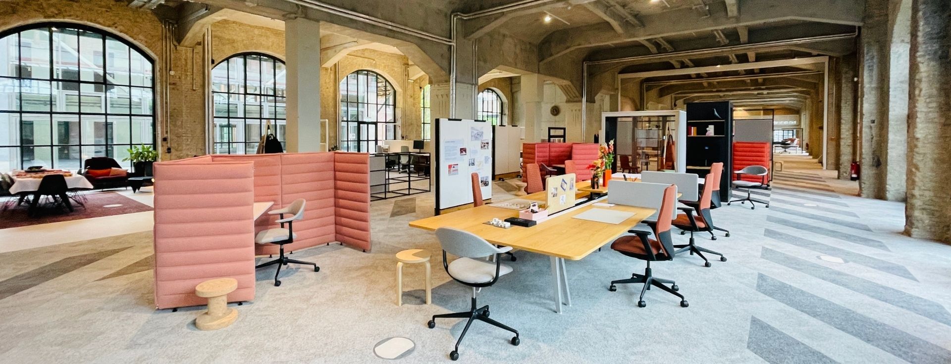 Offene Musterfläche im Vitra BETA Event Space mit verschiedenen Arbeits- und Besprechungsbereichen, abgeschirmten Sitznischen und flexiblen Arbeitsplätzen in historischer Industriearchitektur.