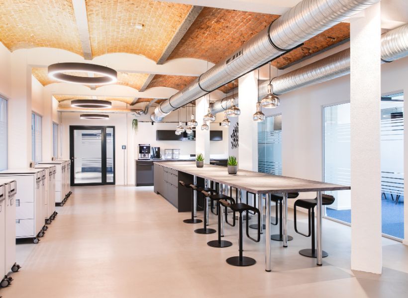 Workcafé mit langer Gemeinschaftstafel, Barhockern und Küchenzeile unter Gewölbedecke als Bereich für Pausen, Austausch und informelles Arbeiten.