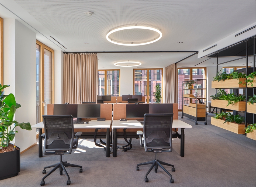 Open-Space-Arbeitsbereich mit integrierter Begrünung durch Pflanzregale, großzügigem Tageslicht und zonierter Struktur für ruhige Arbeitsbereiche.