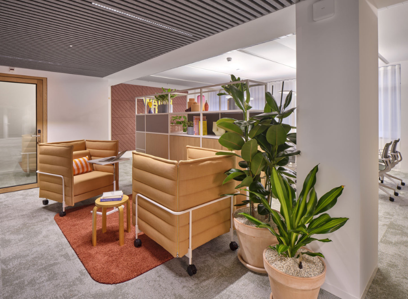 Gemütliche Büro-Loungezone mit gepolsterten Sesseln, Pflanzen, offenem Regalsystem und angrenzendem Konferenzraum.