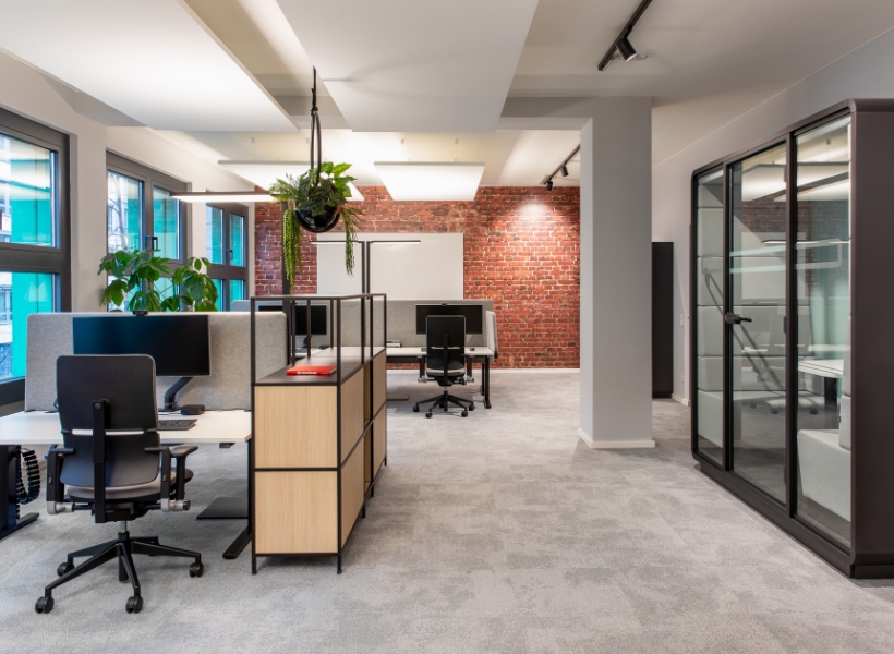 Offene Arbeitslandschaft mit ergonomischen Arbeitsplätzen, Pflanzen und einer verglasten Telefonbox, ergänzt durch eine Backsteinakzentwand.