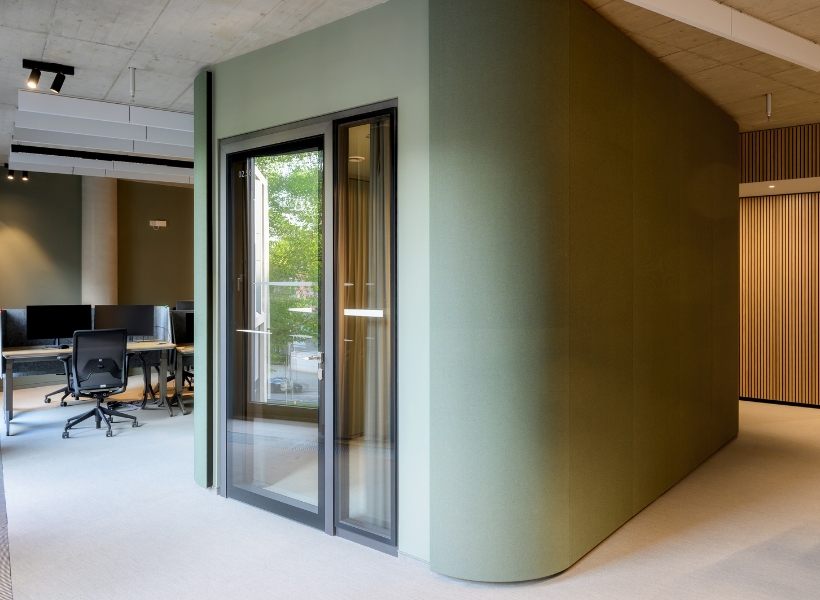 Autarke Raum-in-Raum-Kabine in sanftem Grün, integriert in eine offene Bürofläche mit Arbeitsplätzen und natürlichem Lichteinfall.