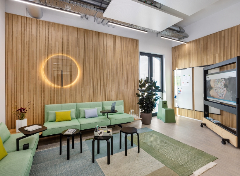 Arbeitslounge mit Sofas, mobilen Tischen, Whiteboards und großem Bildschirm für Besprechungen oder Teamarbeit vor einer Holzlamellenwand.
